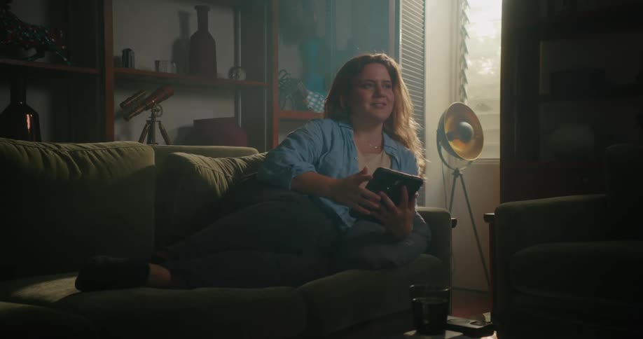 Young happy woman reclined on a couch in the cozy linving room smiles while browsing on a tablet