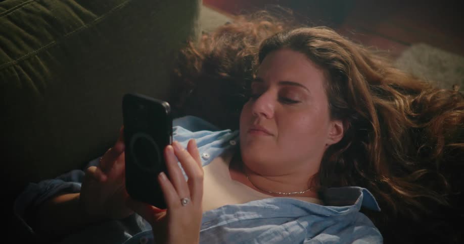 A woman is lying on the living room couch holding her cell phone with both hands as she looks at it
