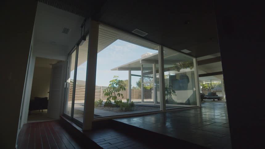 A man looks out a window in his house and walks to another window and looks out