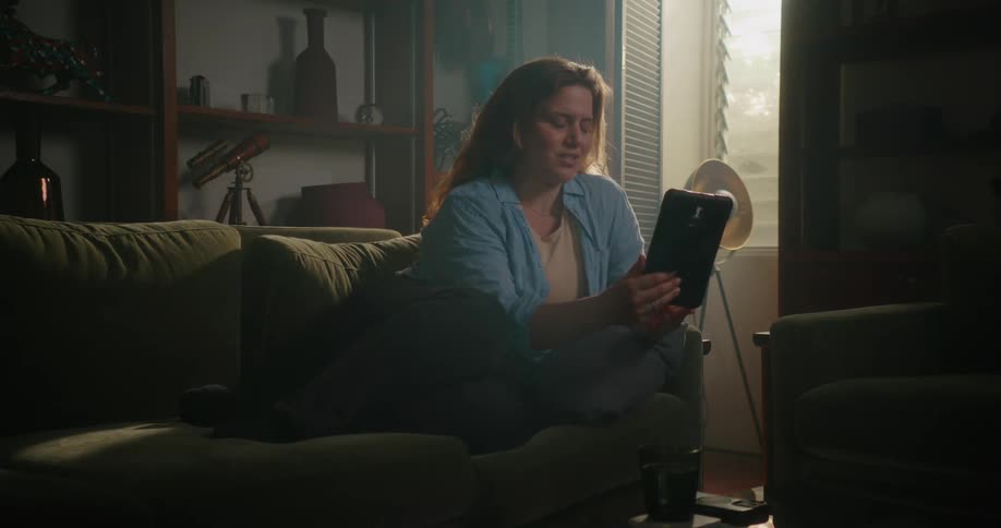Woman with brown wavy hair blue shirt black pants sits on couch making a video call on tablet in the living room