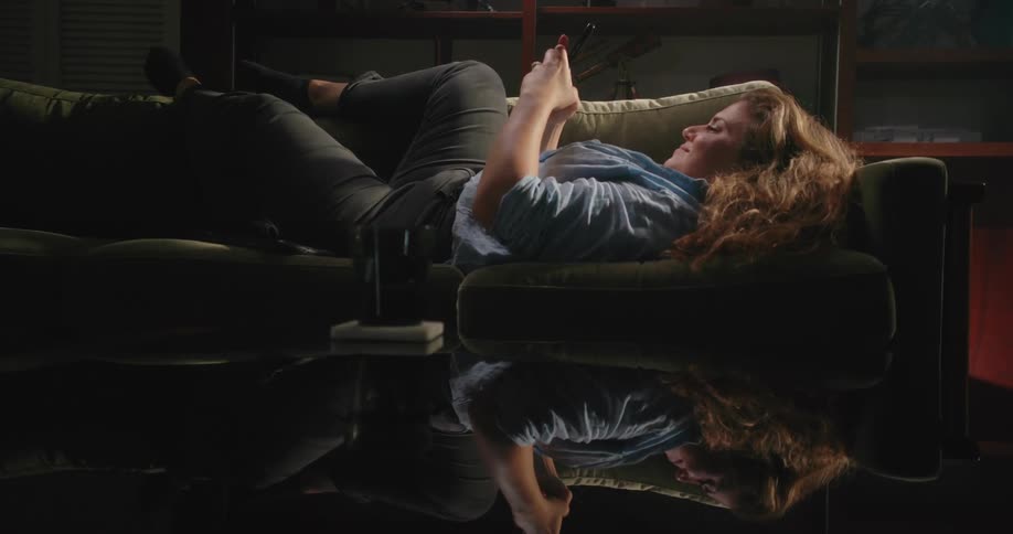 Coffee table surface reflects smiling woman lying down on couch as she texts on smartphone