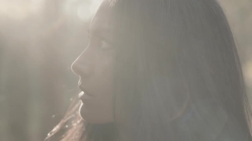 Woman standing in a wooded area looking and turning towards the sun