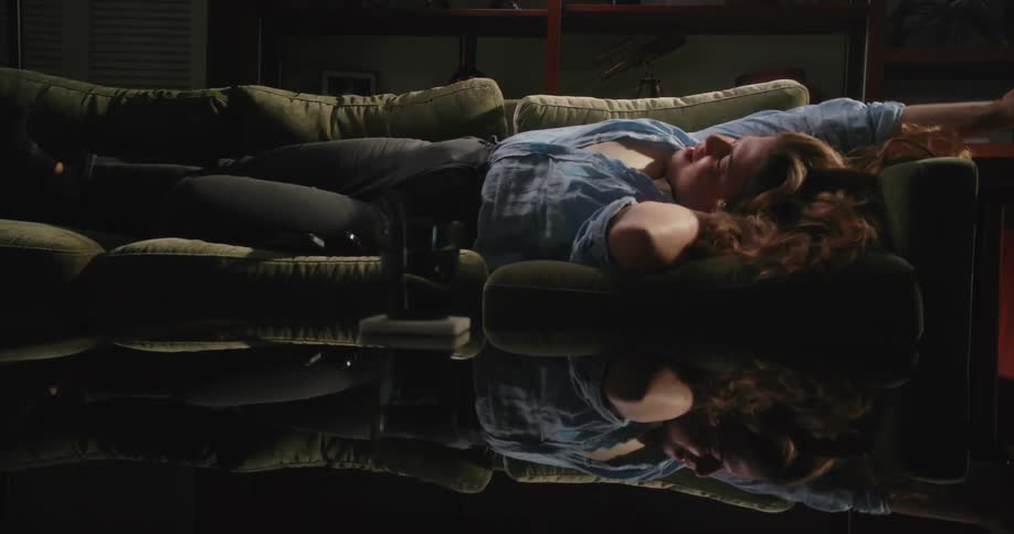Woman with brown wavy hair blue shirt and black pants sleeps on couch yawning and stretching in living room at home