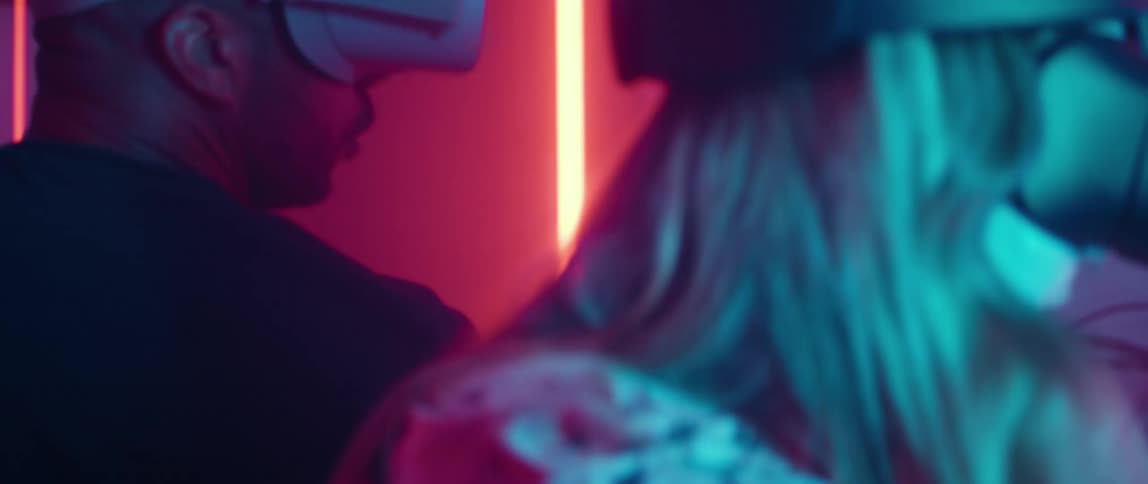 A man and a woman wearing virtual reality headsets have their backs to each other as they play a game in a room illuminated with colored lights