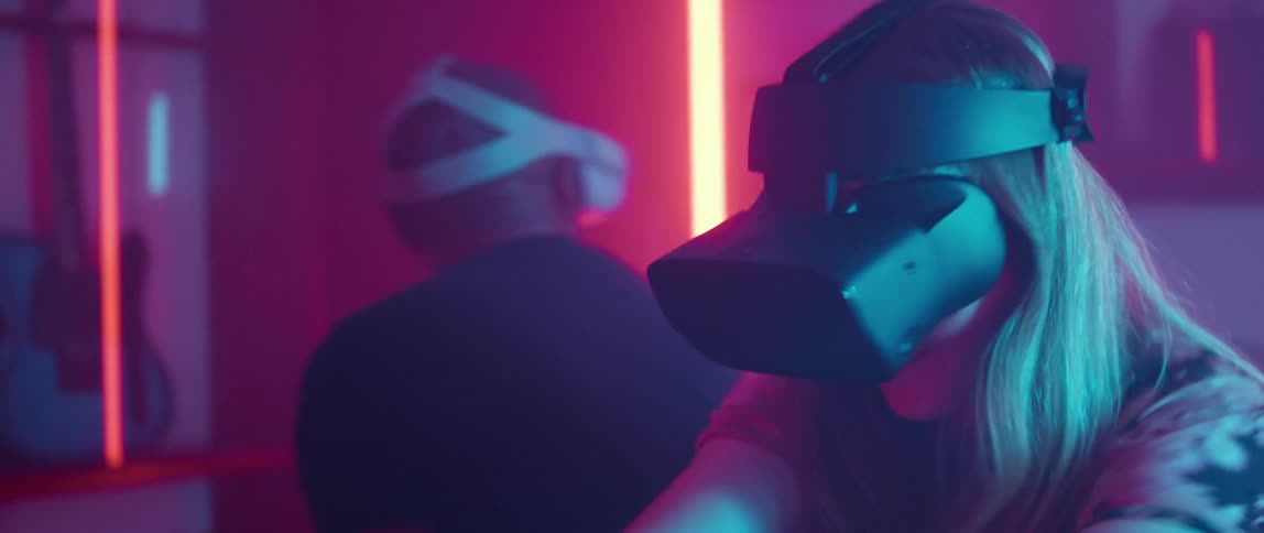 A man and woman laugh as they look around a room and put their hands up in the air as they play a virtual reality game