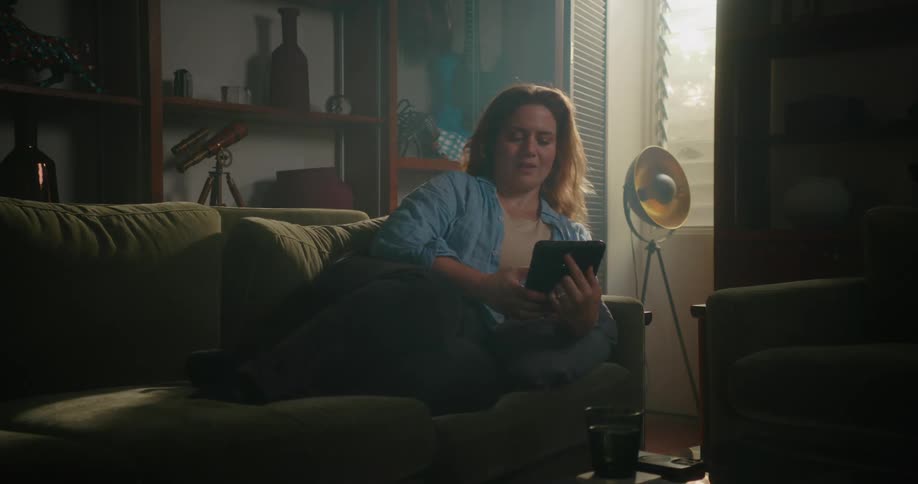 A woman is sitting on the couch in her living room relaxing as she holds a computer tablet