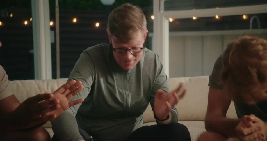 Three men sit on a couch inside a house and one man talks and writes as the other two men high five one another