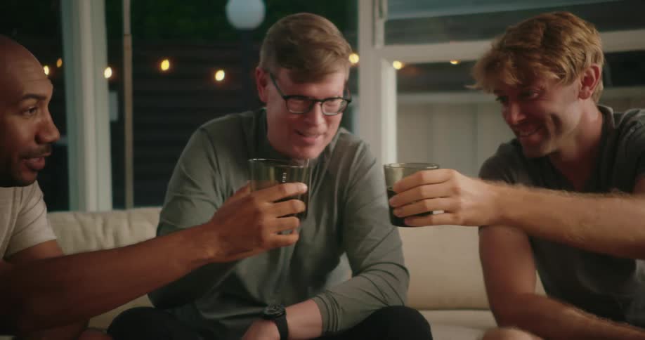 Three men seated on a couch all smile and raise their glasses before drinking