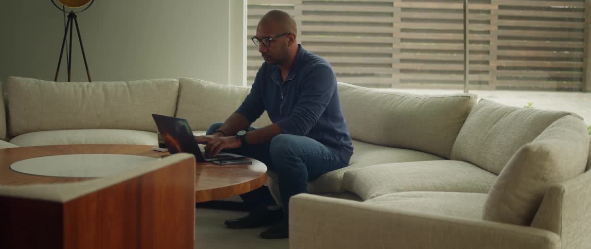 Bald man with glasses blue shirt and jeans sits on sofa using laptop in living room