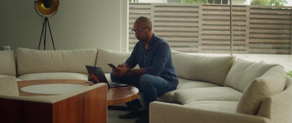 A man is sitting on a couch with a laptop computer on the table as he uses his cell phone