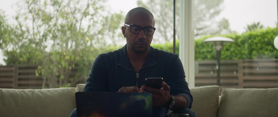 A man is sitting on a couch with a laptop computer on the table as he uses his cell phone