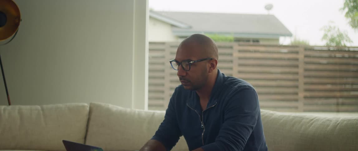 Bald man wearing glasses blue sweatshirt and jeans sits on couch working on laptop in living room