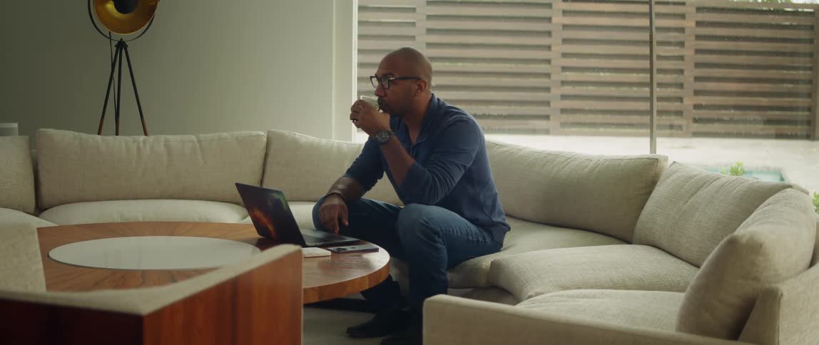 A man sits on his couch and sips a drink while looking down at his laptop