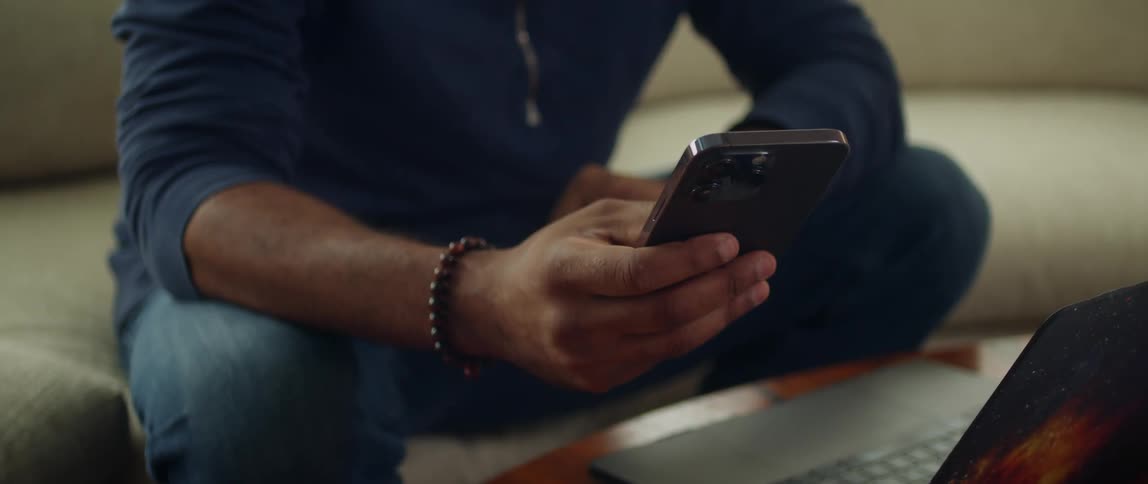 Man with blue sweatshirt and jeans sits on brown sofa using smartphone