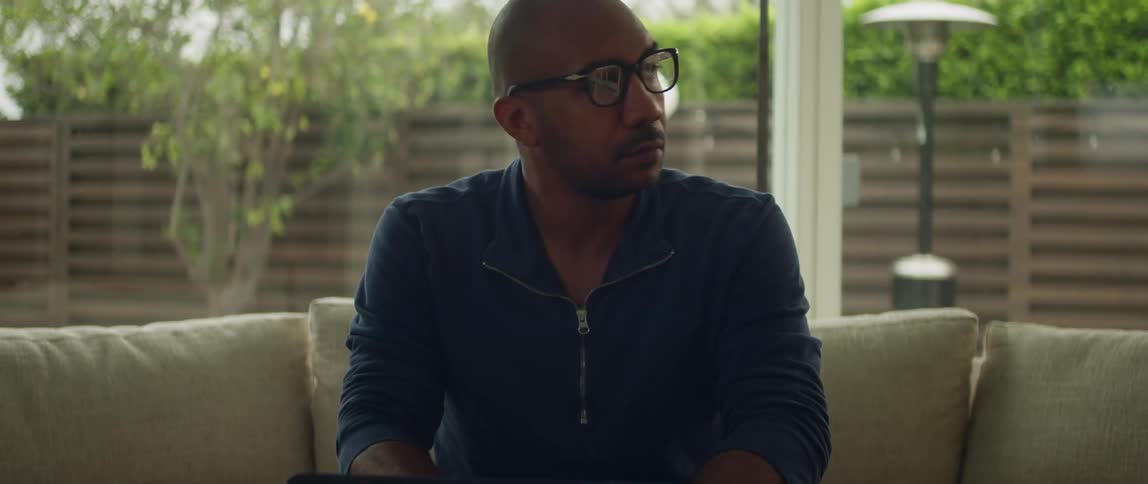 A man is sitting on a couch typing on a laptop computer on the table in front of him as he pauses to think before looking back down at the computer
