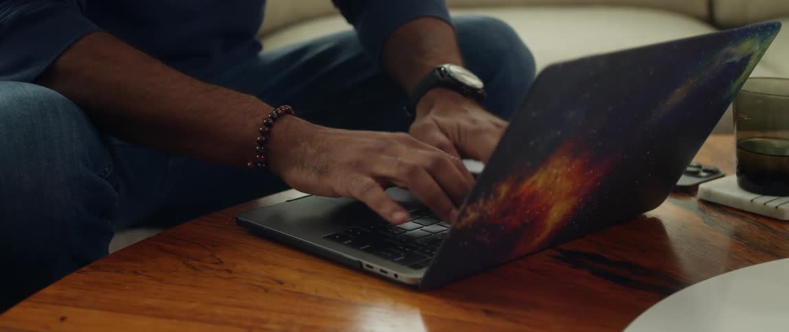 A man sits on a couch and types on a laptop on a table beside a cell phone and a glass of water