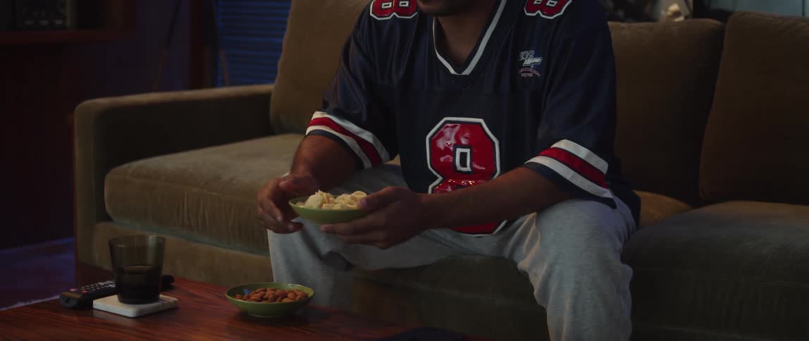 Man wearing sports jersey sits on couch watching while eating snacks