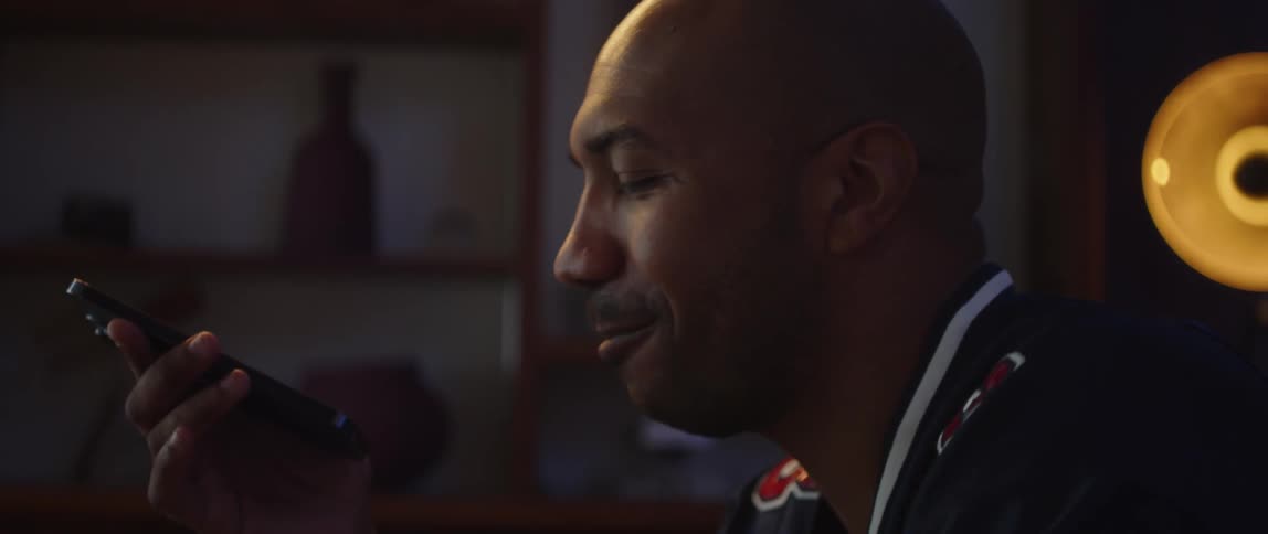 A man is sitting down holding his cell phone as he speaks with a smile on his face