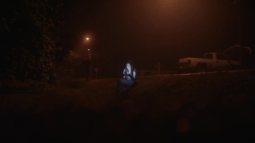 A woman is sitting alone outside on a neighborhood street in the darkness as the light from the cell phone in her hands illuminates her face