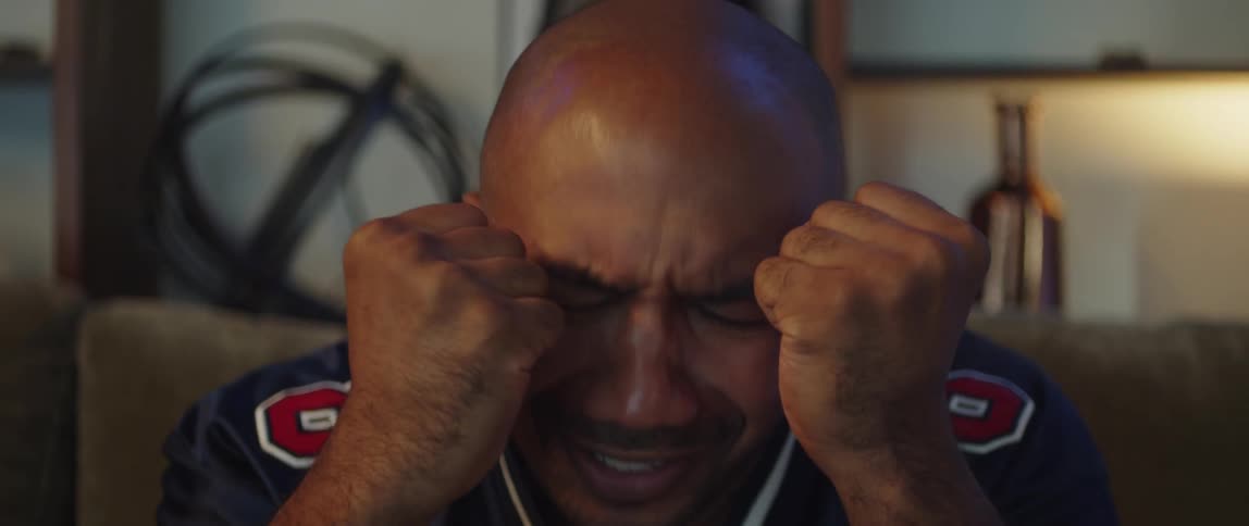 A man sits on a couch and clasps his hands together and shakes his head as he watches something unfold in front of him