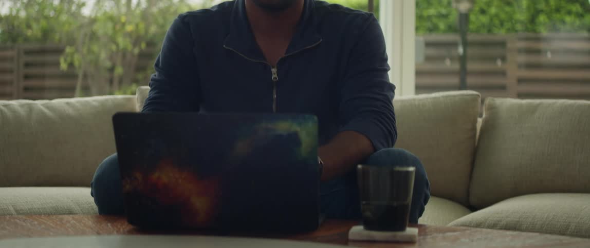 Bald man wearing glasses blue shirt and jeans sits on beige sofa using laptop in the living room