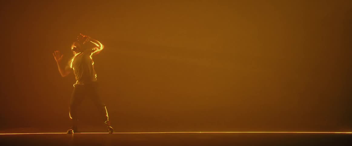 A man is standing in an orange haze moving his arms and dancing with a yellow light shining on him like lightning as he moves