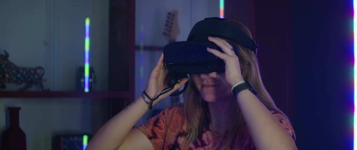A teenage girl is holding on to a virtual reality headset on her head as she listens to someone speak to her and then she pulls it down over her eyes