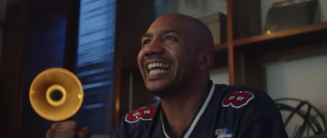 A man wearing a sports jersey is sitting on a couch watching television and he cheers and raises his clenched fists in celebration