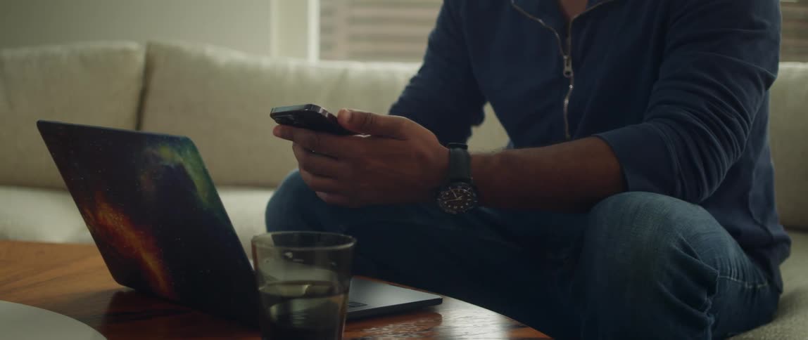 A man is sitting on a couch with a laptop computer on the table in front of him as he holds his cell phone and sets it down next to the computer