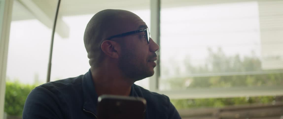 A man is sitting down holding his cell phone and looking around as he speaks with a smile on his face
