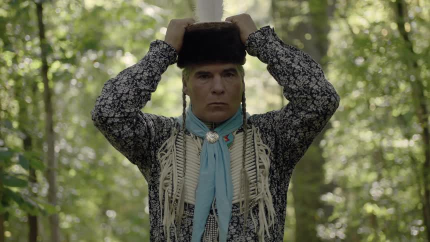 An american indian puts on a fur hat with a feather in it while standing in a grove of trees