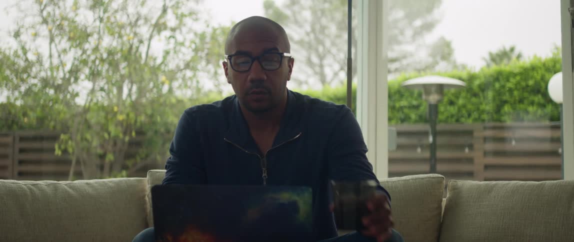Man sits on couch near large windows and takes drink while using a laptop