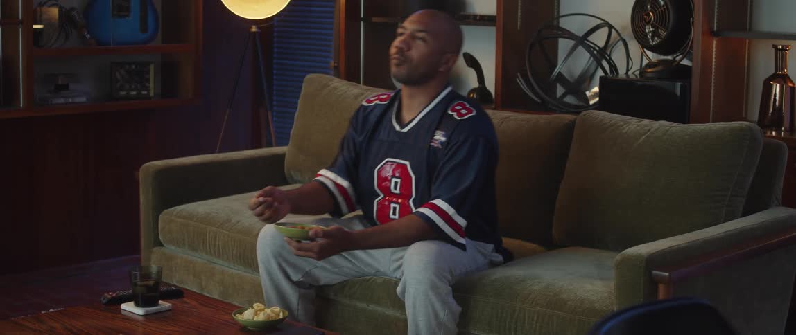Man sits on couch in sports jersey eating snacks while watching game