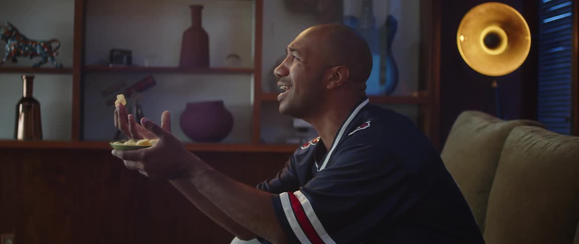 A man in a jersey cheering for his sports team while eating chips on a sofa