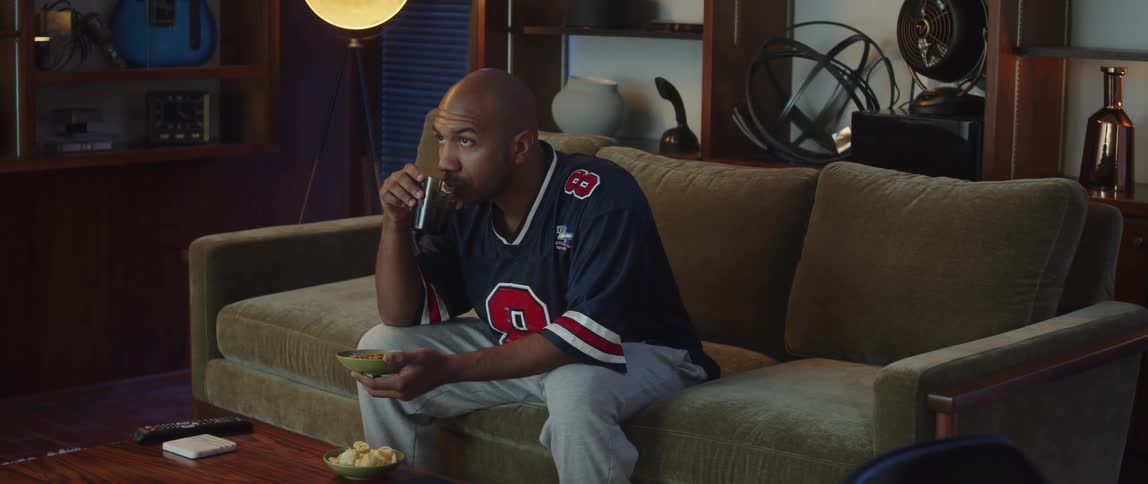 A man wearing a sports jersey is sitting on a couch watching television and holding a bowl of snacks he is eating with his drink
