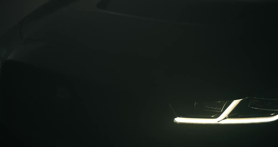 Close up of the headlights of a luxury black car parked on a dimly lit room surrounded by smoke