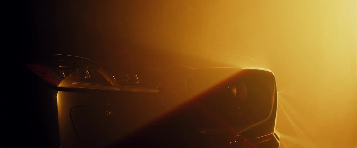 The front of an automobile is seen in a yellow and orange haze with a light above illuminating the grill and headlight
