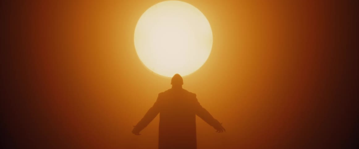 A giant glowing sunlike orb rises over a silhouetted man wearing a trenchcoat