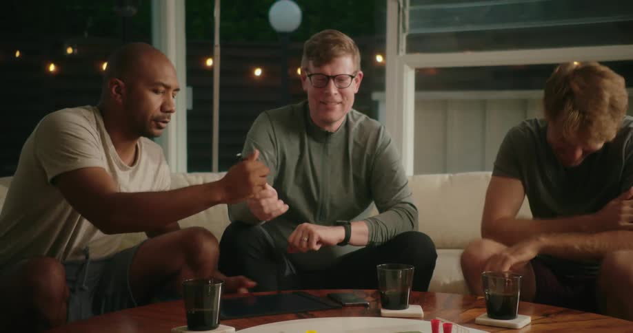 A group of men sit on a couch in a home and turn a table with drinks on it as they laugh and draw on a tablet