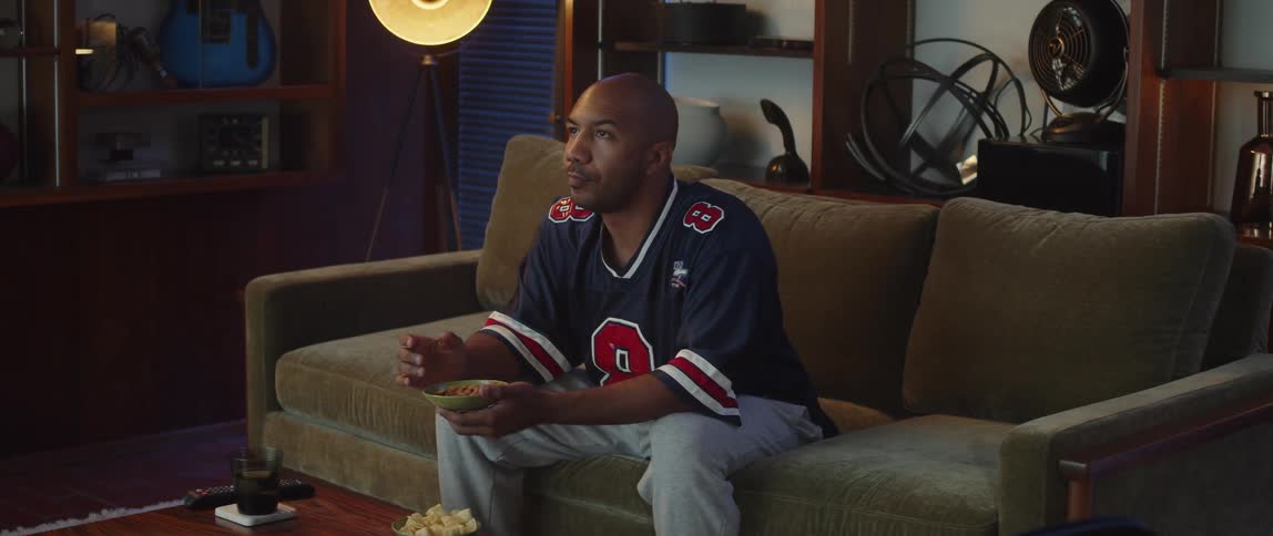 Young man sitting on a couch at home eating snacks watches sports on the television