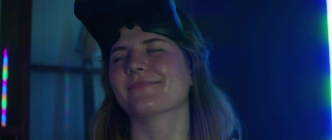 A teenage girl with a virtual reality headset on her head is smiling and shaking her head as she stands in a room illuminated with colored lights