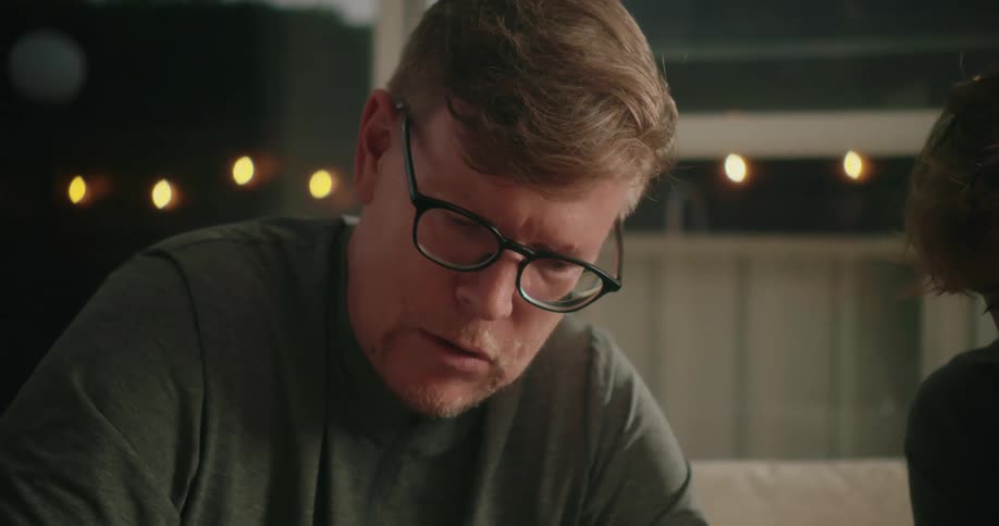 A man sits on a couch at home and talks with a person next to him