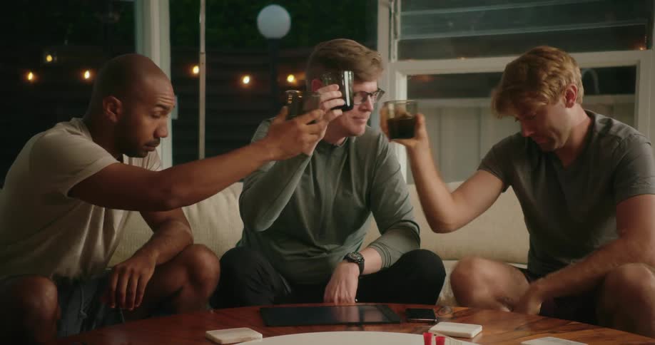 Three men are sitting on a living room couch raising their glasses and touching them as they toast