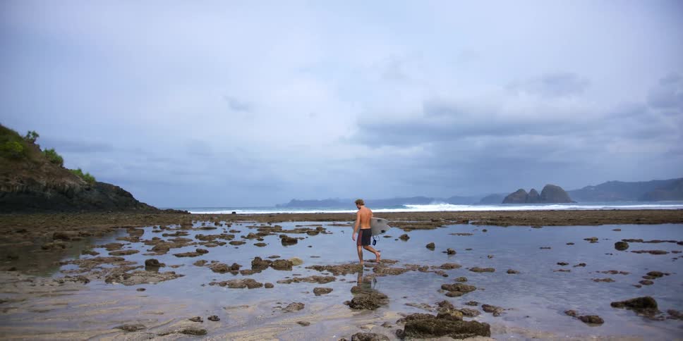 Surfer carrying surfboard picks their way across water on beach as waves crash with foggy mountains in the distance