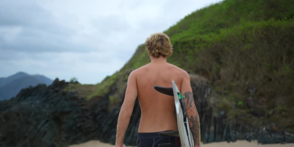 Blonde man with tattoo and shorts carries surfboard on the beach