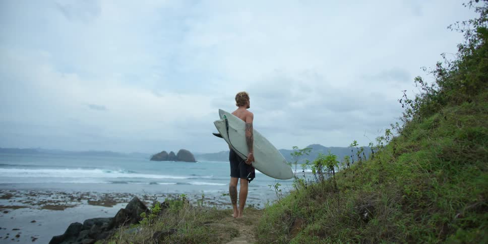 A man carrying a surfboard is walking on a path along the hillside to the beach as waves roll in under a cloudy sky