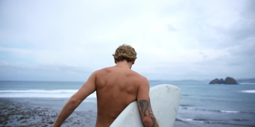 A man carrying a surfboard is walking along the beach as waves roll in under a cloudy sky