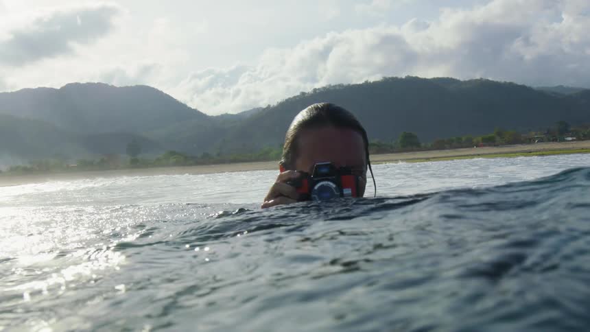 A man standing in a lake with water up to his neck raises a camera and takes a photo