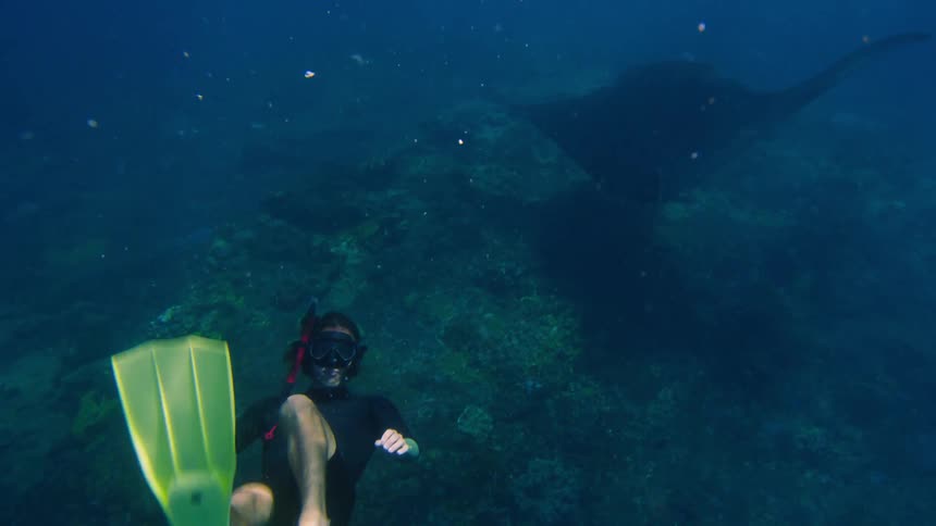 A scuba diver is at the bottom of the ocean swimming facing the camera with a large stingray behind her