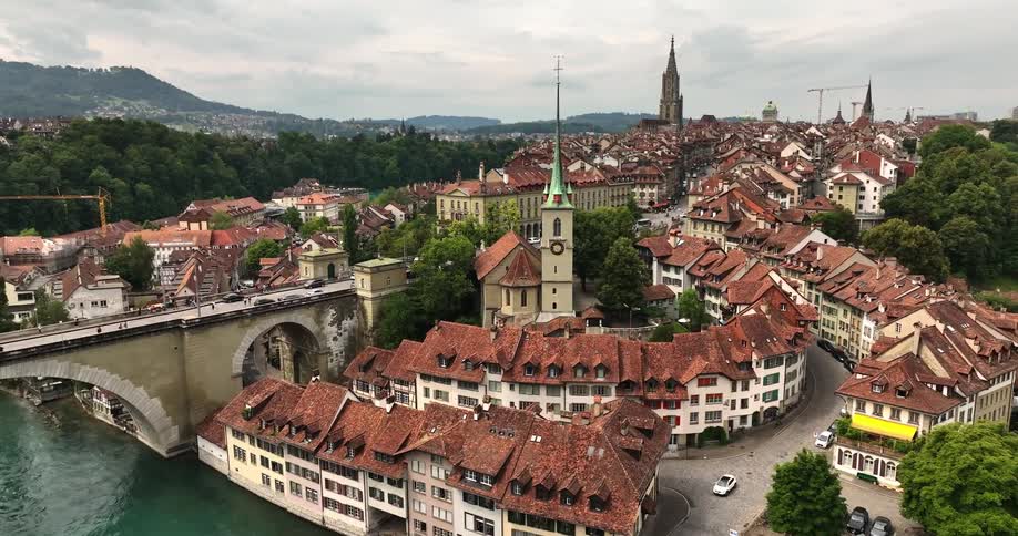 Pedestrians and vehicles cross two bridges in red roofed town with multiple churches as fog covers mountains in the distance